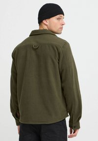 Chaqueta de forro polar verde oliva con cuello, mangas largas y dobladillo redondeado. Presenta un detalle de asa en la parte trasera y puños con botón. Gorro de canale negro.