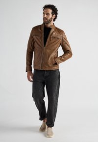 Mann in brauner Lederjacke, schwarzem Hemd, schwarzen Jeans und beigen Slippern, steht mit einer Hand in der Tasche vor weißem Hintergrund.