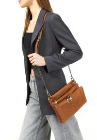 Sac en cuir marron à bandoulière avec deux compartiments zippés, finition texturée et une chaîne réglable, associé à un blazer gris et un jean.