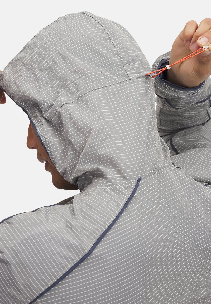 Veste grise texturée avec une capuche, présentant des rayures horizontales blanches, un cordon de serrage réglable et des accents contrastants en bleu marine.