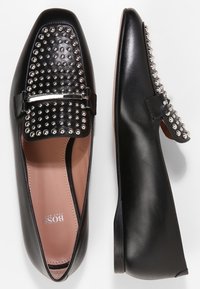 Svarta slip-on skor med en slät läderytan. Ovandelen har silver nitar och ett metallackent rem. Platt häl och rund tådesign.