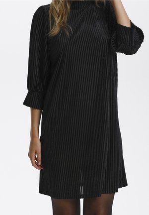 Femme portant une robe noire à côtes verticales, manches longues, avec un col haut et des manches légèrement froncées, sur un fond uni.