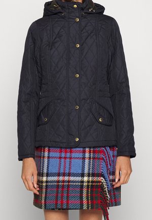 Vrouw draagt een zwarte doorgestikte capuchonjas met gouden knopen over een kleurrijke tartan geruite rok met rode, blauwe en zwarte patronen.