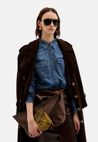 Un manteau en velours côtelé marron, une chemise en denim et une jupe brillante marron. Un grand sac en daim marron avec un portefeuille à motif serpent attacé. Des lunettes de soleil vertes.