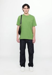 Green cotton t-shirt with a chest pocket, paired with dark blue jeans. Model wears a black shoulder bag and white sneakers.