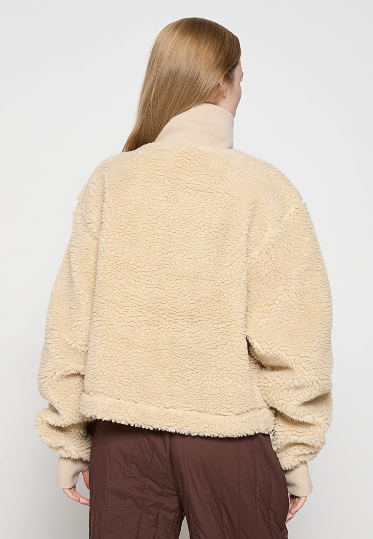 Chaqueta de sherpa beige difusa con un cuello alto, corte oversize y puños elásticos. Tejido texturizado con costuras visibles en el dobladillo y las costuras.
