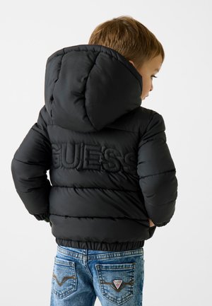 Czarna kurtka puchowa z kapturem, z uniesionym logo „GUESS” na plecach. W komplecie z niebieskimi dżinsami, z widocznymi detalami na tylnej kieszeni.