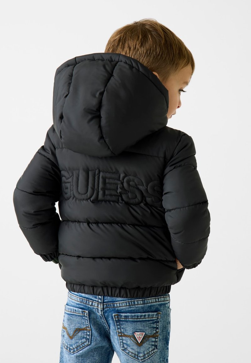 Schwarze Daunenjacke mit Kapuze, mit einem erhabenen "GUESS"-Logo auf dem Rücken. Kombiniert mit blauen Jeans, die Details an der Gesäßtasche zeigen.