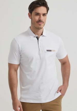 Jeune homme portant un polo blanc à manches courtes avec un petit détail multicolore sur la poche et un pantalon marron, debout avec une main dans la poche.