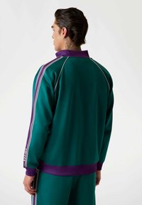 Zelena sportska jakna s visokim ovratnikom, ljubičastim detaljima i bijelim prugama na ramenima. Glatka tekstura i rebrasti rukavi.