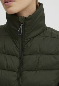 Grön vadderad jacka med hög krage, har en framdragkedja och texturerad quiltad design. Materialet verkar glatt och lätt.