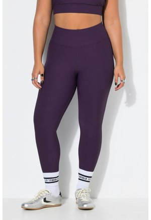 Femme portant des leggings violets taille haute avec un crop top assorti, des chaussettes blanches avec texte noir et des chaussures de sport argentées, debout contre un mur blanc.