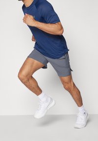 Camiseta deportiva azul marino, pantalones cortos grises y zapatillas blancas. Hombre demuestra una postura de carrera, mostrando material transpirable y ajuste cómodo.
