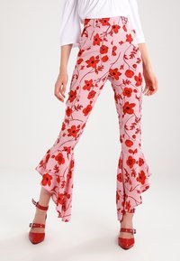 Bloemenprint broek met een lichtroze achtergrond, gekenmerkt door rode bloemen en klokvormige ruffles aan de zoom. De stof lijkt licht en soepel.