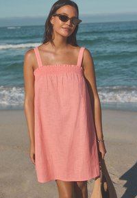 Rosa linnetklänning med en smockad överdel och breda band. Bärs på en strand, kombinerad med solglasögon och en flätad väska. Enkel, luftig design.