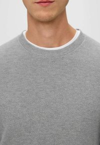 Grauer Strickpullover mit strukturiertem Muster und Rundhalsausschnitt, getragen über einem weißen Unterhemd. Nahaufnahme des Brustbereichs.