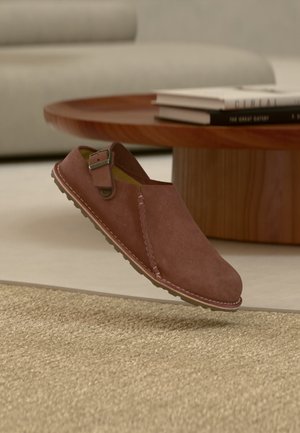 Brązowe zamszowe buty wsuwane z paskiem na klamrę, widocznym przeszyciem, leżące obok drewnianego okrągłego stołu z ułożonymi książkami w pomieszczeniu.