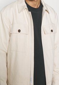 Veste en coton beige avec une fermeture éclair à l'avant, deux poches poitrine et des boutons foncés. Portée sur une chemise noire. Tissu texturé, design structuré.