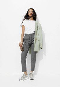 T-shirt blanc, blazer vert clair, pantalon gris ajusté et baskets argentées. Tenant une pochette au motif léopard. Textures lisses, design décontracté.