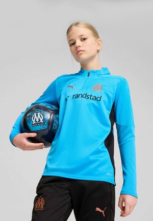 Giovane calciatrice con la maglia blu del Marsiglia che tiene una palla ufficiale del Marsiglia su uno sfondo semplice.