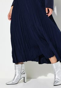 Stivali d'argento alla caviglia con punta a punta e tacco a blocco abbinati a un vestito pieghettato blu navy. La texture del vestito è fluida e morbida.