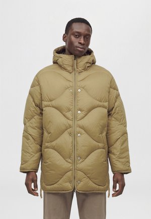 Un homme porte un manteau matelassé beige à capuche, avec une fermeture éclair sur le devant et des boutons-pression, sur un fond clair uni.