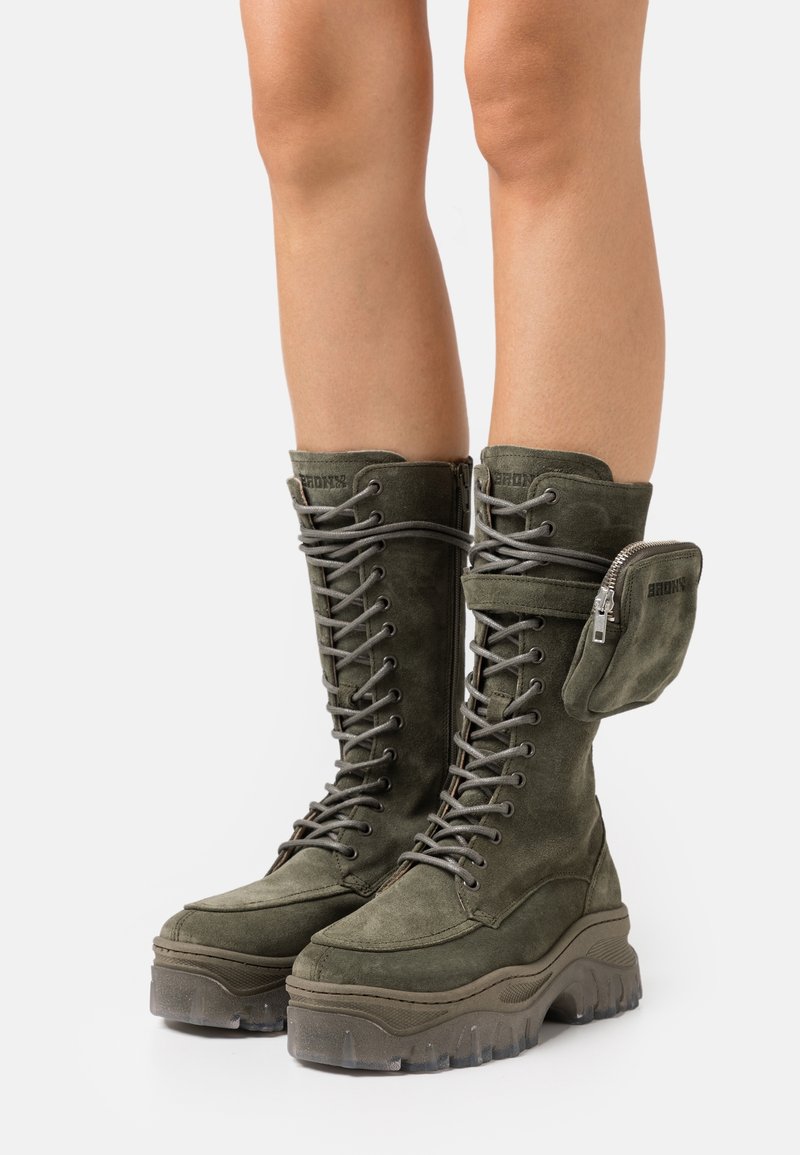 Bottes kaki en daim montant jusqu'au genou avec semelles épaisses, lacets à l'avant, fermeture éclair latérale et une pochette zippée amovible sur le mollet extérieur.
