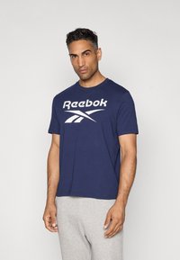 Navy blue cotton t-shirt featuring a white Reebok logo, crew neck, short sleeves, and a relaxed fit. Worn with grey sweatpants.