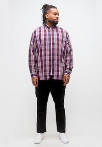 Ανδρας με dreadlocks που φοράει πουκάμισο καρό κόκκινο, άσπρο και μπλε, μαύρο παντελόνι και λευκά αθλητικά παπούτσια, στέκεται μπροστά σε ένα απλό λευκό φόντο.