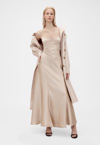 Robe beige en satin avec un corsage ajusté et une jupe fluide, associée à un trench-coat beige clair doté de gros boutons et d'une coupe décontractée.