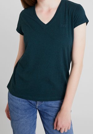 Femme portant un T-shirt vert foncé à col en V et manches courtes ainsi qu’un jean bleu, debout, une main détendue le long du corps.