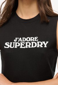 Donna che indossa un top senza maniche nero con la scritta bianca "J'ADORE SUPERDRY" sul petto, con le braccia incrociate in vita.