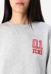 Szürke pulóver kerek nyakkivágással, piros hímzett "01" és "ICHI" felirattal. Puha, textúrált anyagból készült.