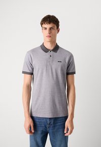 Γκρι πόλο μπλουζάκι με διακοσμητική �υφή, κοντά μανίκια και σκούρο γιακά. Έχει μικρό λογότυπο στο στήθος. Φοράει μπλε τζιν.