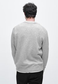 Szary sweter dziany z okrągłym dekoltem i ściągaczami na rękawach. Materiał wydaje się miękki i lekko teksturowany. Widoczny od tyłu, noszony na białej koszuli.