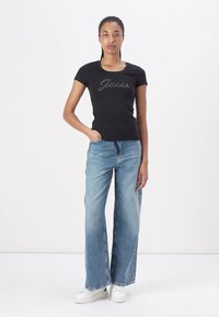 Black short-sleeve t-shirt with "Guess" logo in rhinestones, paired with light blue wide-leg denim jeans and white sneakers.