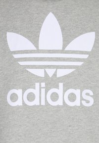 Camisa de algodón gris con un gran logo trefoil blanco de Adidas que presenta tres hojas sobre rayas horizontales y la palabra "adidas" debajo.