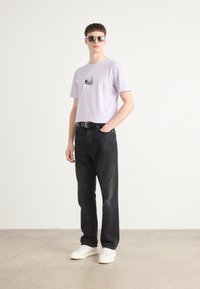 Lilac cotton t-shirt with a graphic print, paired with black denim pants and white sneakers. Model wearing reflective sunglasses.