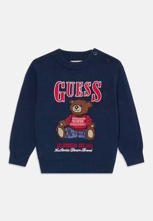 Marineblå barnekjole med rød "GUESS" tekst, en kosebamse iført en rød genser, og broderi av "Los Angeles EST. 1981".
