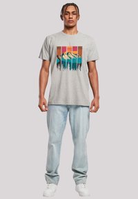 Grijze katoenen t-shirt met een kleurrijke berg- en bosafbeelding. Model draagt lichtblauwe jeans en witte sneakers. Casual pasvorm.