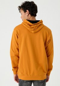 Sudadera con capucha naranja de corte relajado, que presenta un cordón en la capucha y puños acanalados. La tela parece suave con una textura lisa.