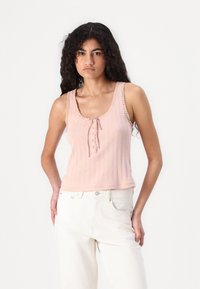 Light pink tank top with ribbed texture, a scoop neckline, decorative lace trim, and a tie front; worn with white high-waisted pants.