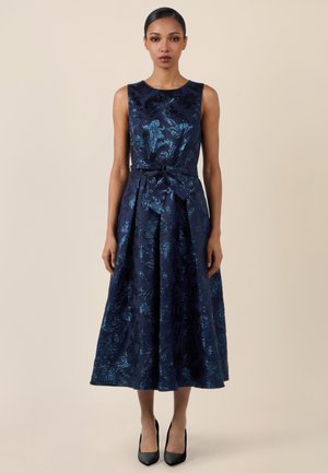 Frau in einem ärmellosen, marineblauen Midikleid mit Blumenmuster und Taillengürtel, steht aufrecht vor einem einfarbigen beigen Hintergrund.