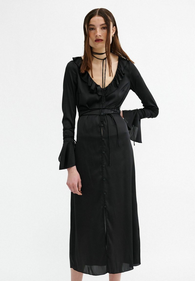 Schwarzes Satin-Kleid mit Rüschendetail entlang des Ausschnitts, langen Ärmeln mit Glocken-Manschetten und einer Frontknopfleiste. Mit Taillenteil zum Binden.
