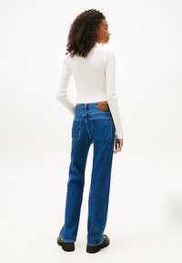 Mujer con cabello rizado que lleva una blusa blanca de manga larga, vaqueros de tiro alto azul y zapatos negros, de pie de espaldas contra un fondo blanco.