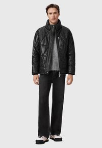 Sort quiltet puffjakke lavet af skinnende materiale, med høj krave og lynlåslukning, båret over en grå sweater og sorte jeans.