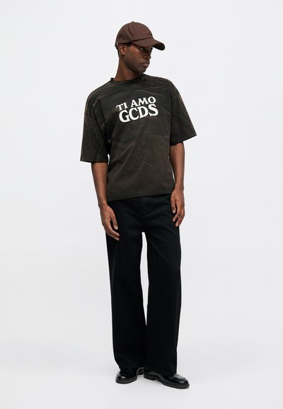 Svart oversized t-skjorte med hvit tekst "TI AMO GCDS," kombinert med vide svarte bukser og en brun caps, med en glatt tekstur.