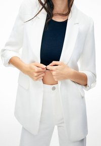 Blazer blanco ajustado con puños enrollados, combinado con una blusa de punto azul marino ajustada. Textura suave y diseño estructurado. Pantalones blancos con detalles de botones.