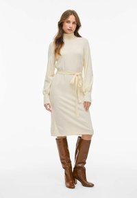 Robe en tricot crème avec de longues manches bouffantes, un col haut et une ceinture à nouer assortie à la taille, associée à des bottes marron hautes jusqu'aux genoux.