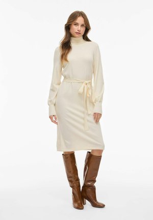 Robe en tricot crème avec de longues manches bouffantes, un col haut et une ceinture à nouer assortie à la taille, associée à des bottes marron hautes jusqu'aux genoux.